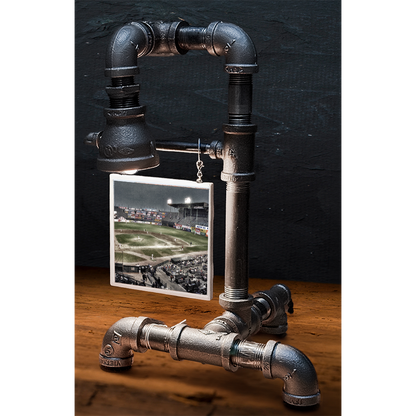 Industrial-style table lamp made from black metal pipes with a small framed baseball stadium photo hanging from the pipe, on a wooden surface