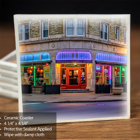 A square ceramic coaster featuring a colorful street photograph of Colter Bay restaurant storefront at night, shown standing on a wooden surface with a blurred background. Text in the lower-left corner reads: 'Ceramic Coaster 4 1/4 x 4 1/4" Protective Sealant Applied Wipe with damp cloth'.
