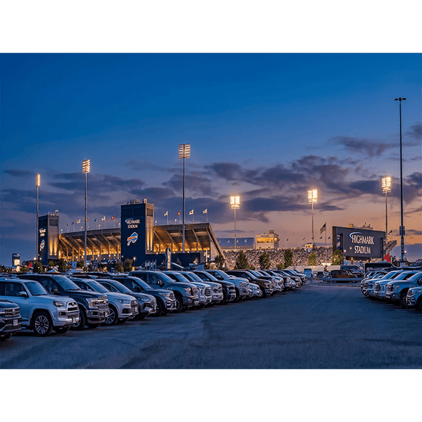 Highmark Stadium at dusk with illuminated floodlights, parked cars in foreground, blue sky with clouds, and flags flying, Buffalo Bills home stadium